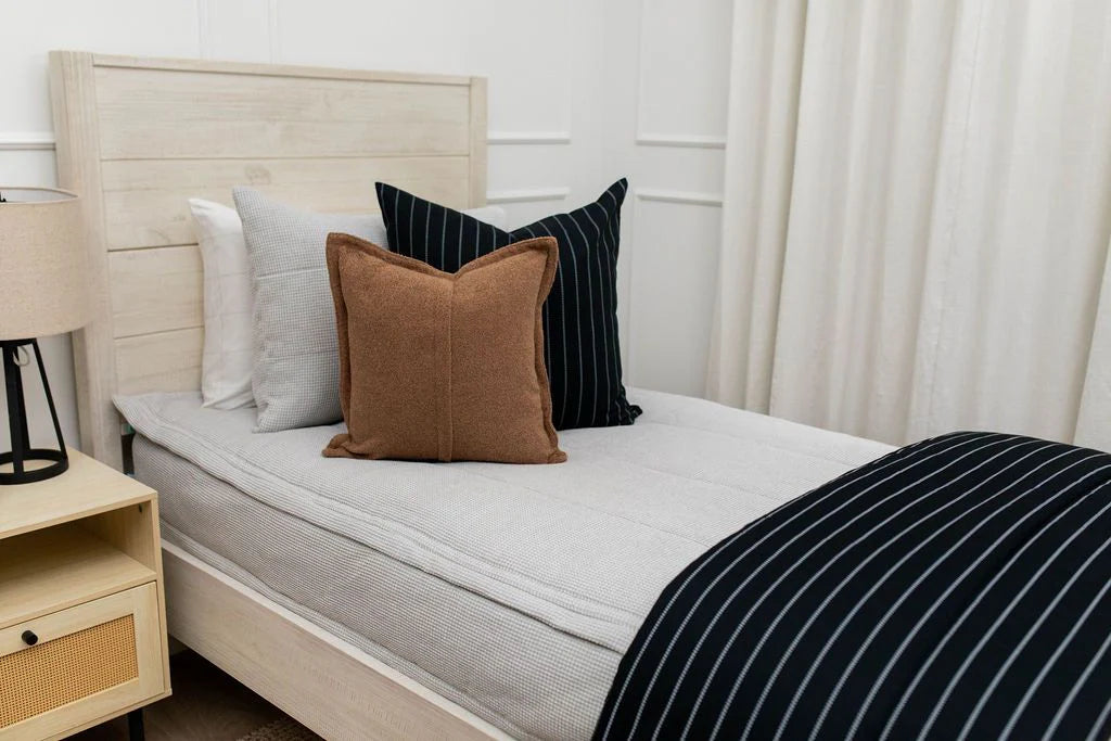 A neatly made single bed with a light wood headboard features black and white striped bedding, a brown accent pillow, and a black striped pillow. The bed is elegantly draped with the Bryant Luxe Blanket by Beddy’s. To the left sits a lamp on a small bedside table, while white curtains hang in the background.