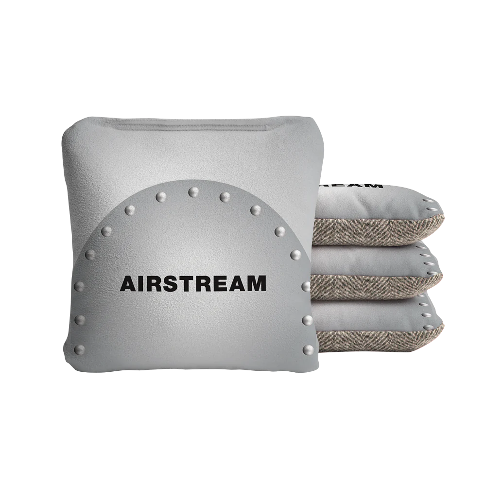Four square pillows—three stacked in tweed fabric and one in front featuring a metallic, riveted design with “AIRSTREAM” in bold black lettering—give the Airstream ACA Cornhole Bags a stylish, personalized flair for your game setup.