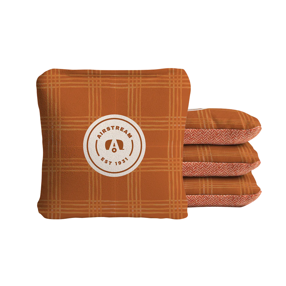 Four orange plaid Airstream ACA Cornhole Bags are stacked; the front bag features a circular logo with “Airstream Est 1931” around a stylized “A.” The other three bags, showcasing a striking chevron pattern reminiscent of custom cornhole designs, are stacked behind the front bag.