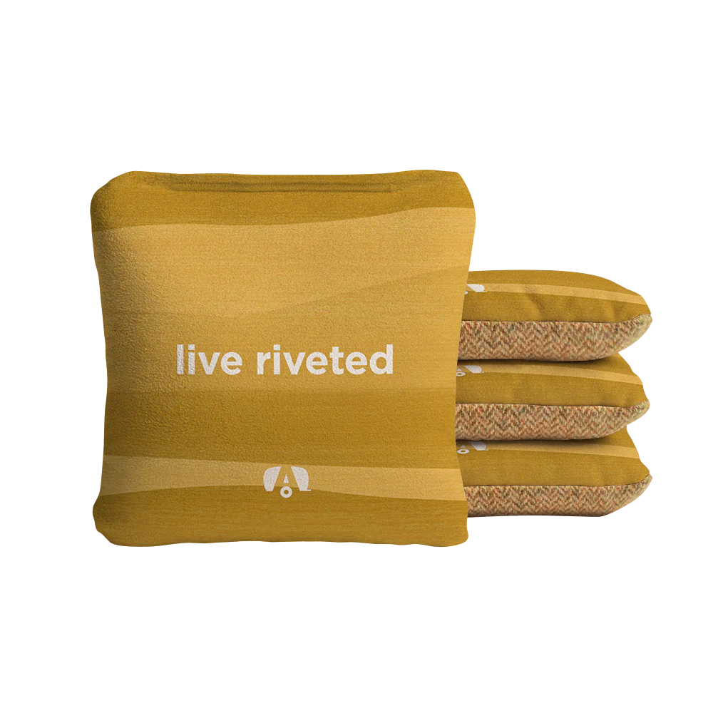 Four Airstream ACA Cornhole Bags in mustard yellow are stacked, with the front one featuring the white “live riveted” text and a small white logo beneath it. The other three bags display a patterned side, reminiscent of custom cornhole bags. The background is plain white.