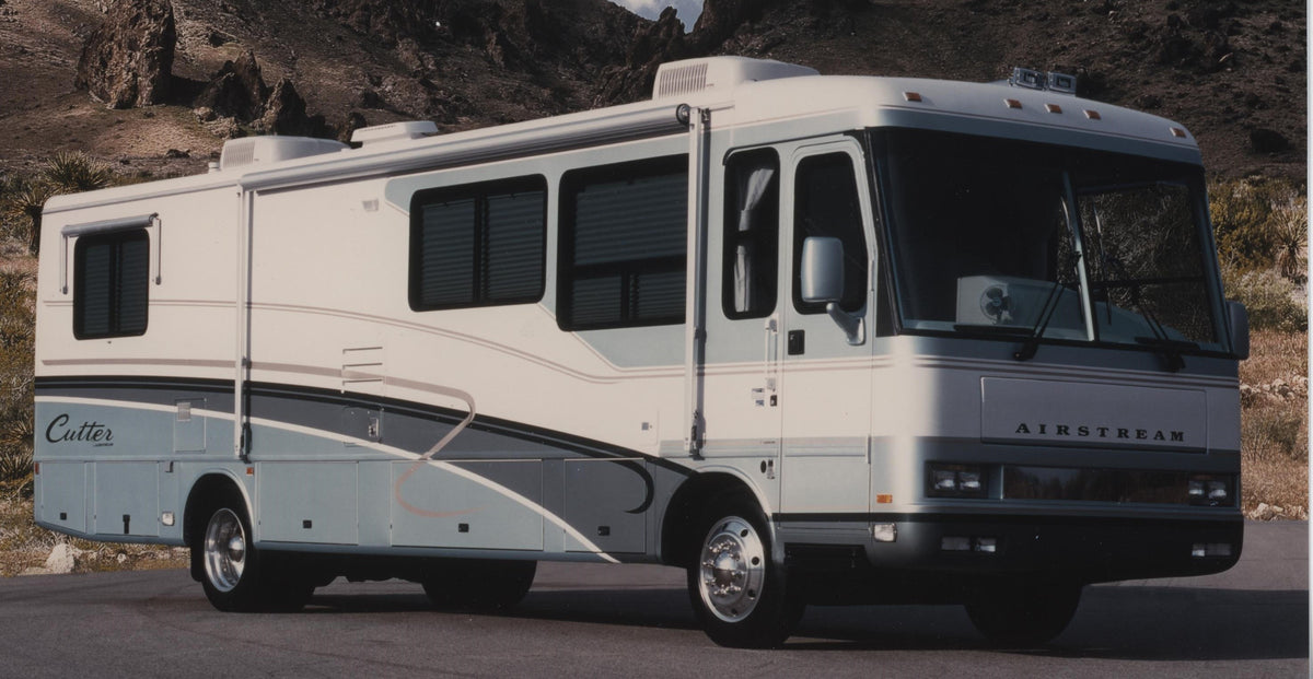 A large white and light blue Airstream Cutter motorhome is parked on a paved road with rocky hills in the background. The vehicle features tinted windows and multiple storage compartments along its side, providing comfort inside and out—enhanced by custom-made bedding that includes luxurious Airstream Custom 200 Thread Count Sheets for Cutter Motorhomes, ensuring restful sleep wherever your journey leads.