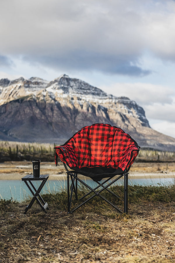 A red and black buffalo plaid camping chair with a black frame is set up outdoors next to a small folding side table with a cup, with a mountain and lake in the background.