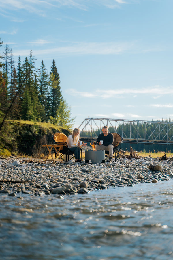 A couple sits by a campfire on a rocky riverbank outdoors, with evergreen trees, a metal bridge, and blue sky in the background.