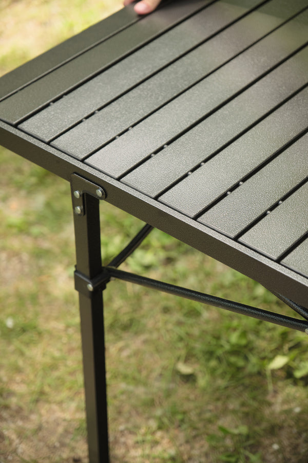 Close-up of a dark gray slatted outdoor table with a black metal leg and bracket, set on grass with a hand resting on the table edge.