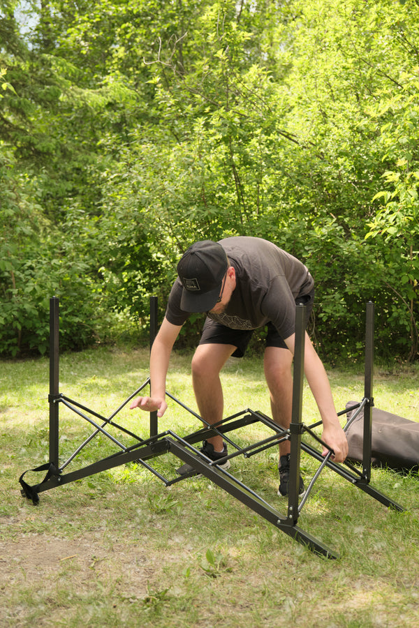 A man in a black t-shirt and shorts is assembling a black metal folding stand outdoors on a grassy lawn with trees in the background.