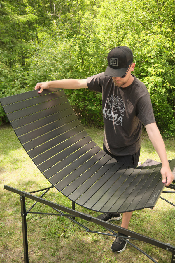 A man outdoors on grass in front of dense green trees is demonstrating a curved black slatted lounge chair or fire pit cover resting on a metal frame. He is wearing a black Kuma t-shirt and a black baseball cap.