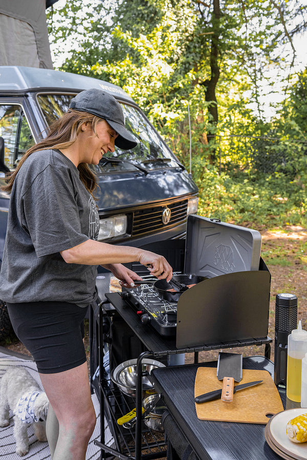A woman wearing a gray t-shirt and black cap cooks at an outdoor mobile kitchen setup next to a dark gray VW camper van, with a dog at her side and a wooded forest background.