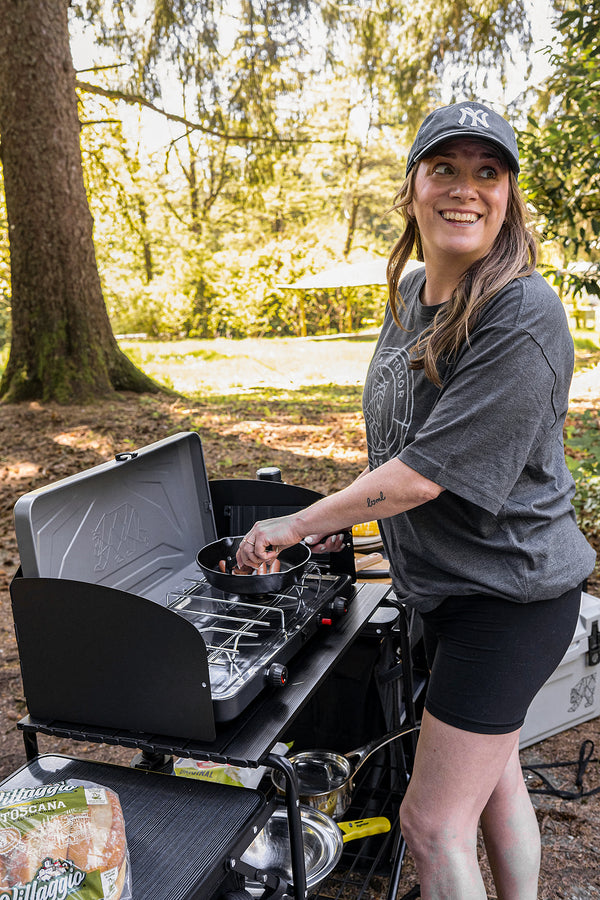 A woman wearing a black baseball cap and a dark gray t-shirt stands next to a black portable camping stove, cooking food in a pan. The setting is outdoors in a wooded area with trees in the background.
