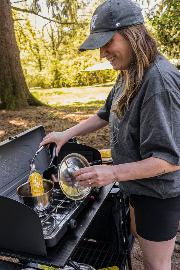 A woman outdoors by a campfire or portable stove, holding a glass lid while another hand holds a pot with a corn cob. She wears a dark gray t-shirt, black shorts, and a dark gray baseball cap.