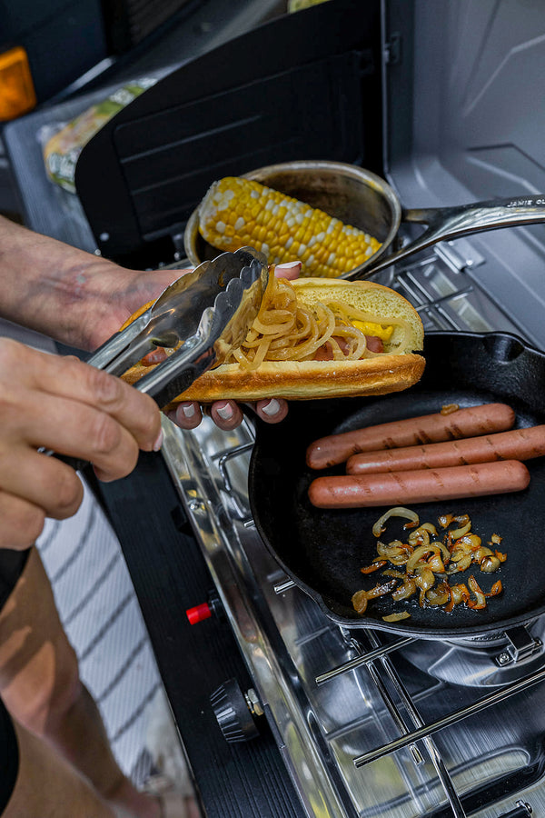 A close-up action shot of a person using metal tongs to assemble a hot dog with caramelized onions, with a cast iron skillet containing hot dogs and onions on a portable stove in the background.