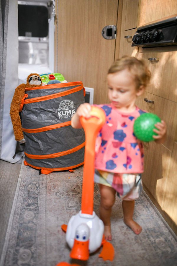 A toddler stands barefoot in a compact camper kitchen area, holding an orange and white duck-shaped toy push toy while also holding a green ball. A gray and orange Kuma Pop-up Play Tent is visible in the background.