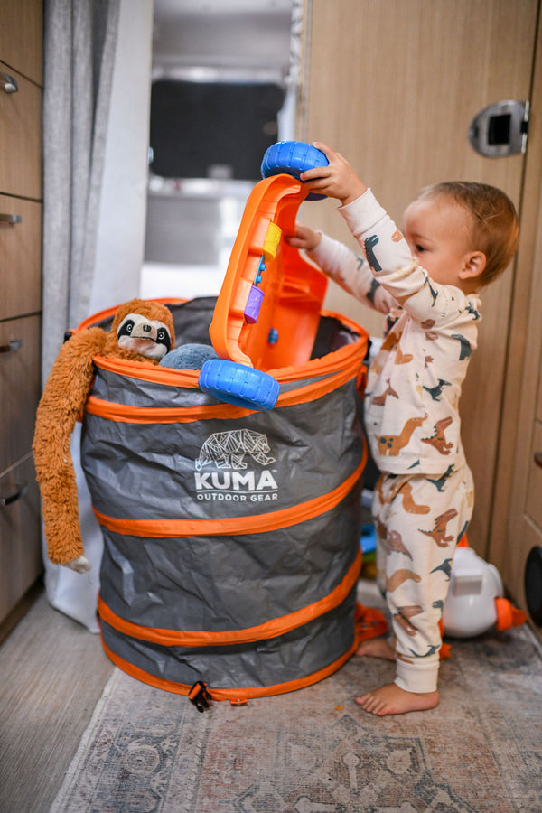 A toddler in dinosaur-print pajamas stands next to a gray and orange Kuma collapsible storage bag, reaching into it for a blue toy. A plush sloth toy is tucked inside the bag, which is set on a patterned rug inside what appears to be an RV or play area.