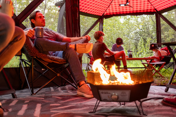 A man relaxes in a camp chair beside a small fire pit with flames burning inside, while children sit nearby under a red canopy in a wooded outdoor setting.