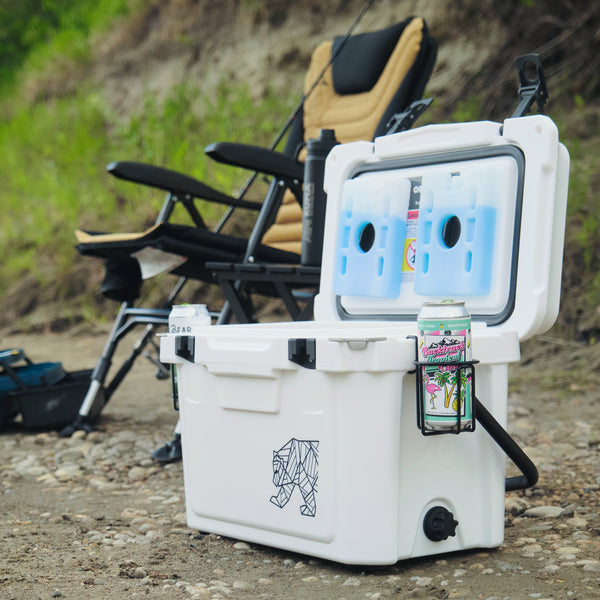 A white cooler with an open lid, shown outdoors on a gravel surface with a camping chair and grassy background. The cooler has a black geometric animal logo on the front, a side handle, and a black cup holder holding a can and small item.