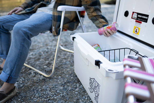 A person sitting in a folding chair beside an open white cooler, holding a pink beverage can. The cooler has a black wire basket and a black line-art animal logo on the side.