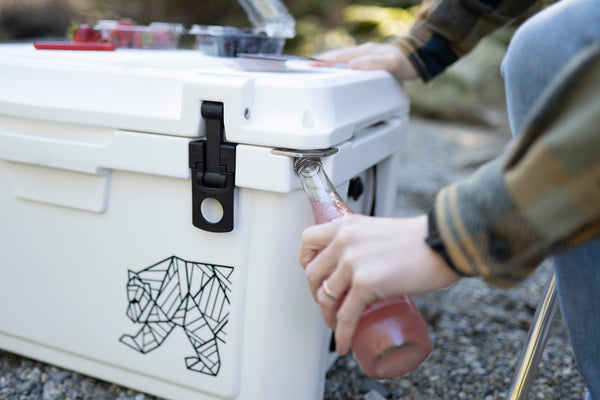 A white cooler with a black geometric animal graphic on the front, shown outdoors with a person pouring a pink drink from a glass bottle into it.