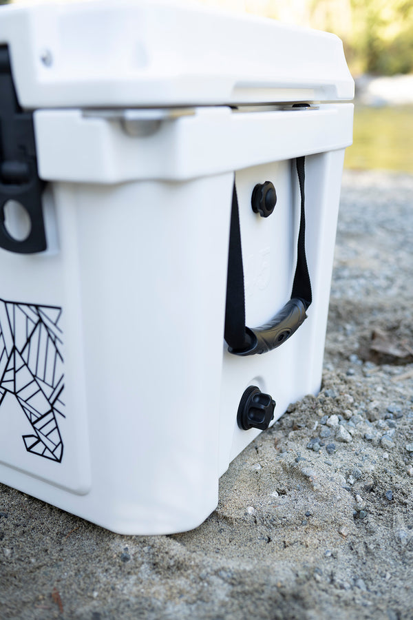 A close-up outdoor shot of a white cooler sitting on gravel near water, showing the side latch, carry handle, and a black geometric graphic print on the front.