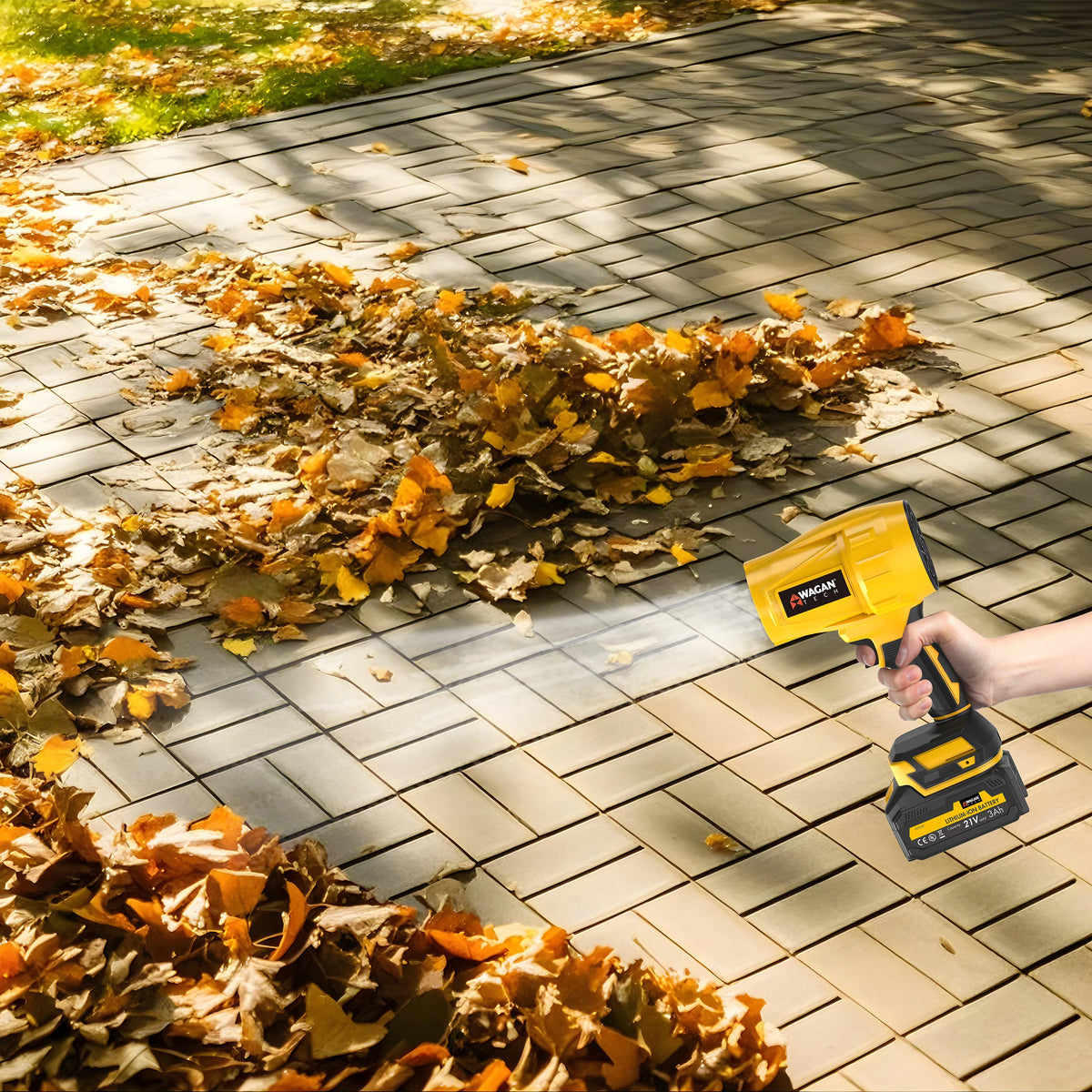A hand uses the Jetblow by Wagan Tech, a yellow compact blower, to clear autumn leaves from a paved outdoor walkway during campsite cleanup as sunlight shines through the trees above.