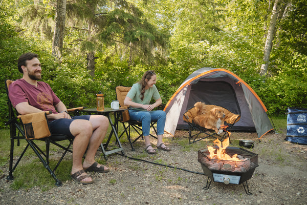 A man and a woman relax in camping chairs beside a fire pit and tent in a wooded outdoor setting. A camping hammock is draped over a folding chair, and a small table holds cups and a bottle between them.