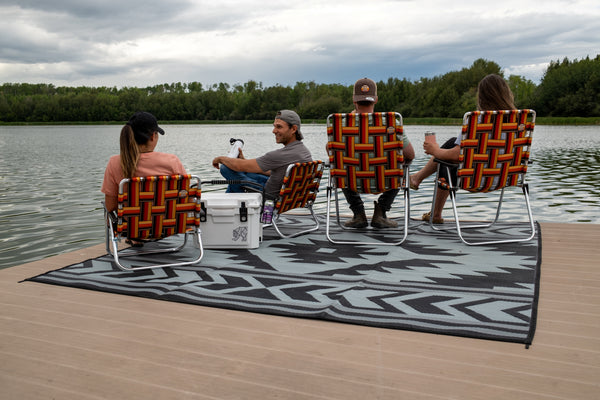 Four people sitting in woven outdoor lounge chairs on a large black, gray, and white patterned rug on a wooden dock by a lake, with trees and an overcast sky in the background.