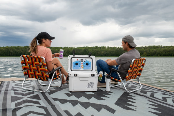 A man and a woman sit in woven camping chairs on a patterned dock, facing each other across a lake with a white portable power station between them. The scene is outdoors under a cloudy sky with trees in the distance.