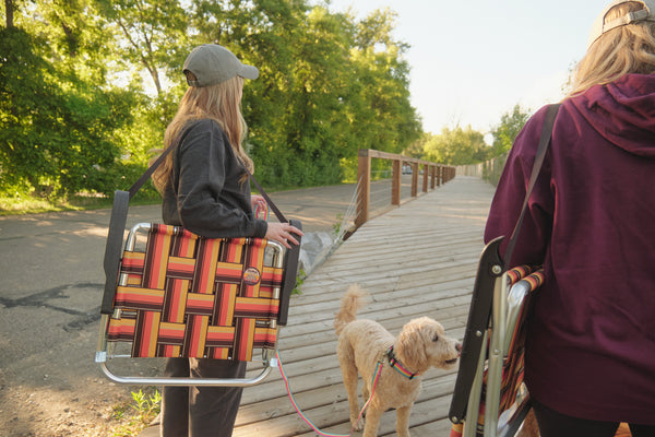 A woman on a wooden boardwalk carries a folding pet stroller with a woven red, orange, and yellow patterned fabric, accompanied by a small light-colored dog. Another person is partially visible on the right.