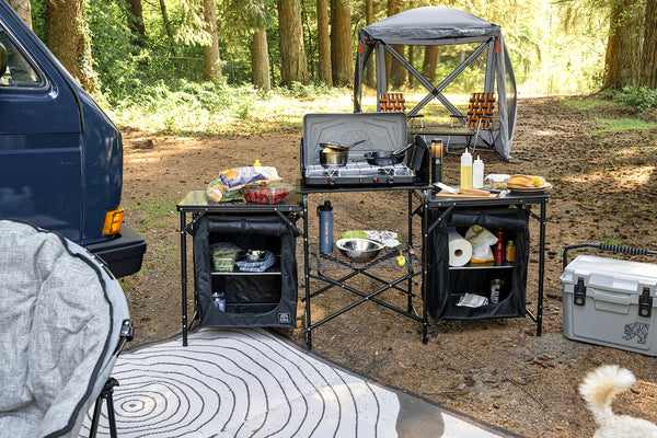 A black folding camp kitchen setup in a forest campsite, featuring a central gas stove with a canopy above, two side storage tables with shelves, cooking accessories, and a cooler nearby. A blue vehicle is partially visible on the left, and a dog is present in the lower right corner.