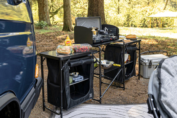 A black folding camp kitchen set up outdoors at a campsite next to a blue vehicle. The set includes a small table with a hanging black storage bag, a cooking pot, a condiment bottle, and a second folding table with shelves and a hanging bag.