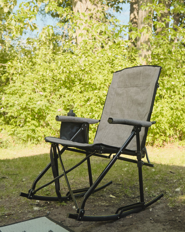 A black and gray folding camping rocking chair with a built-in cup holder, set outdoors on a grassy lawn with a background of green trees.