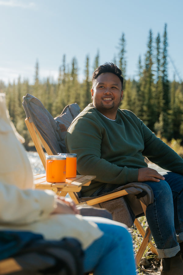A man sitting in a wooden folding chair outdoors by a lake, wearing a green long-sleeve shirt and blue jeans. Two orange drink cups sit on the chair beside him, with pine trees and blue sky in the background.