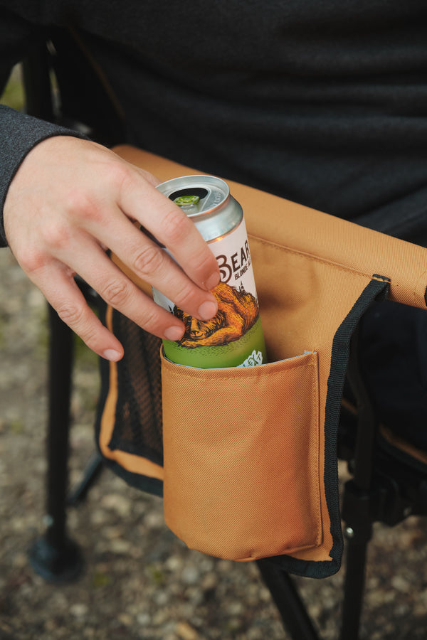A tan canvas beverage holder pocket attached to a black folding chair, holding a green can of beer, with a person's hand reaching in.