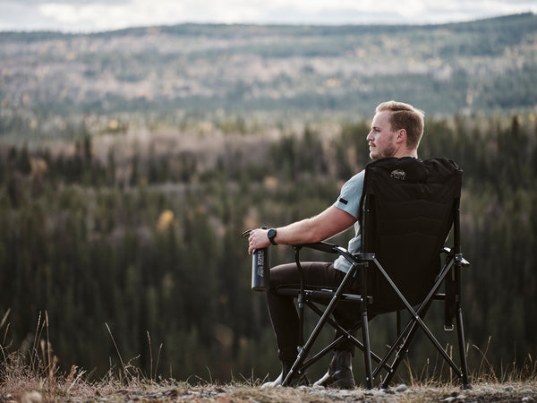 A man sitting in a black camping folding chair on a grassy hill, looking out over a forested mountain landscape.