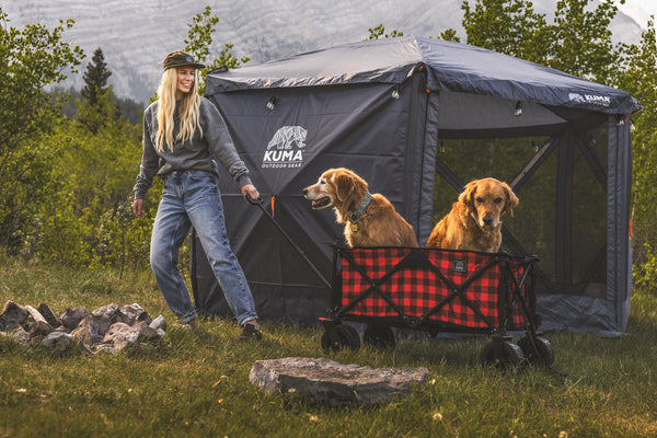 A woman outdoors next to a gray Kuma canopy tent with two golden retriever dogs sitting in a red-and-black plaid Kuma dog wagon. Trees and mountains are visible in the background.