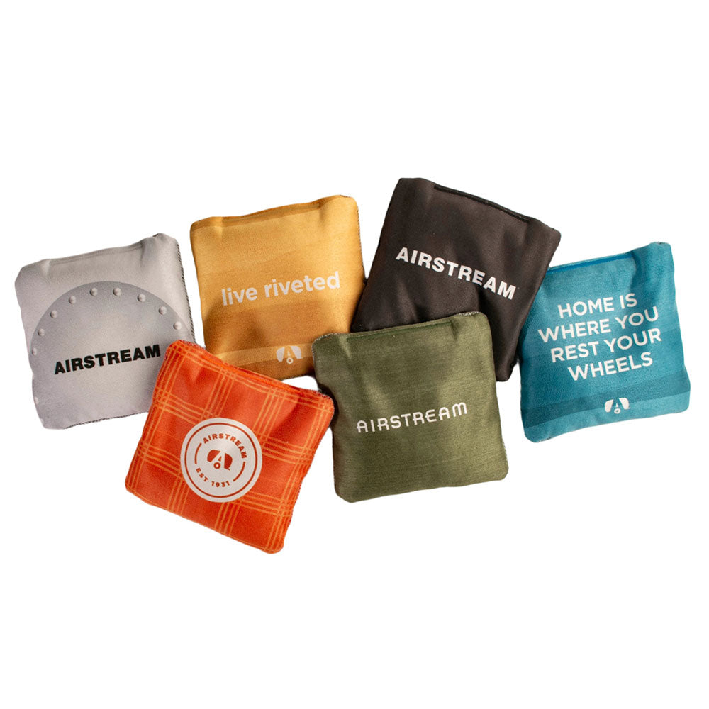 Six vibrant, fabric Airstream ACA Cornhole Bags are displayed together, each showcasing unique Airstream-themed text or graphics—such as “live riveted” and “HOME IS WHERE YOU REST YOUR WHEELS.” Crafted with quality and style, these custom bags are perfect for bringing personality to your next cornhole game.