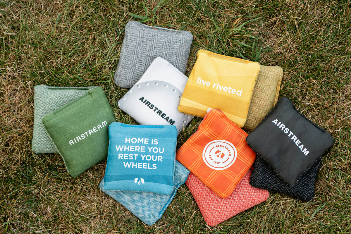 Nine colorful Airstream ACA Cornhole Bags are arranged on grass, featuring custom text such as “Home is where you rest your wheels” and “live riveted.”.