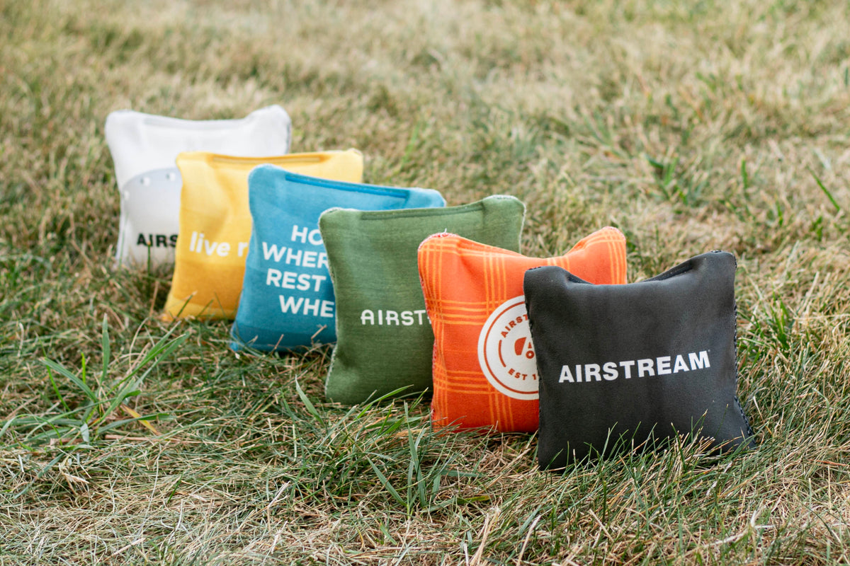 Five Airstream ACA Cornhole Bags, each featuring unique patterns and text, are arranged in a row on dry grass outdoors. The bags are available in white, yellow, blue, green, orange, and black.