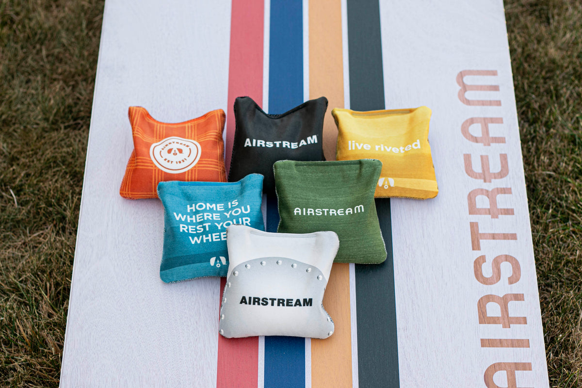 Six vibrant Airstream ACA Cornhole Bags, each featuring unique Airstream-themed designs and slogans, are displayed on a vertically striped cornhole board with “AIRSTREAM” across it, set on a grassy lawn.