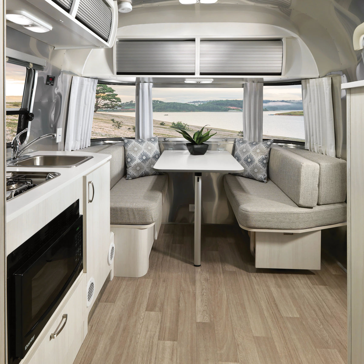 Modern RV interior featuring a compact kitchen with a microwave, sink, and a dining area furnished with cushioned benches and a table. Large windows—beautifully framed by Airstream Solid Custom Curtains for Bambi Travel Trailers—provide stunning views of the lake and surrounding landscape.
