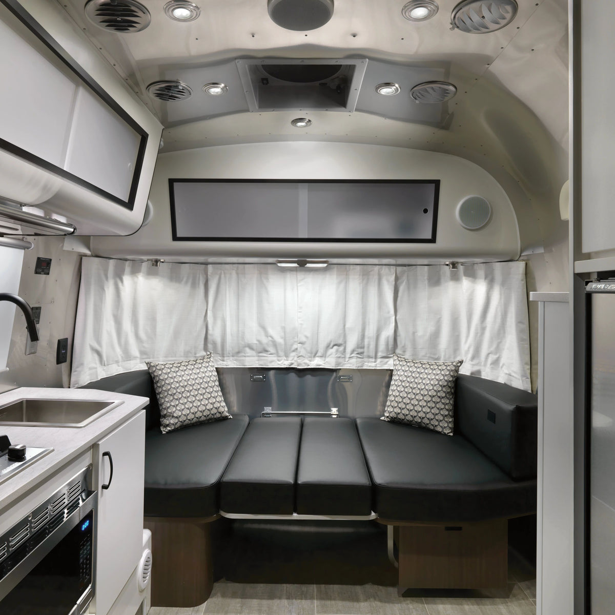 Modern camper van interior with a black cushioned seating area, patterned pillows, Airstream Solid Custom Curtains for Caravel Travel Trailers in crisp white, and a small kitchen area featuring a sink and microwave. Rounded walls and ceiling lights complete the cozy space.