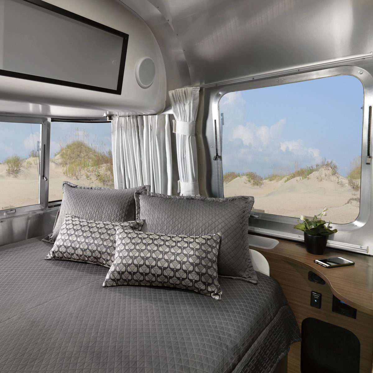A neatly made bed with gray bedding sits inside an Airstream trailer, where large windows are dressed with Airstream Solid Custom Curtains for Caravel Travel Trailers, framing views of sand dunes and a blue sky. A small plant rests on the wooden nightstand.