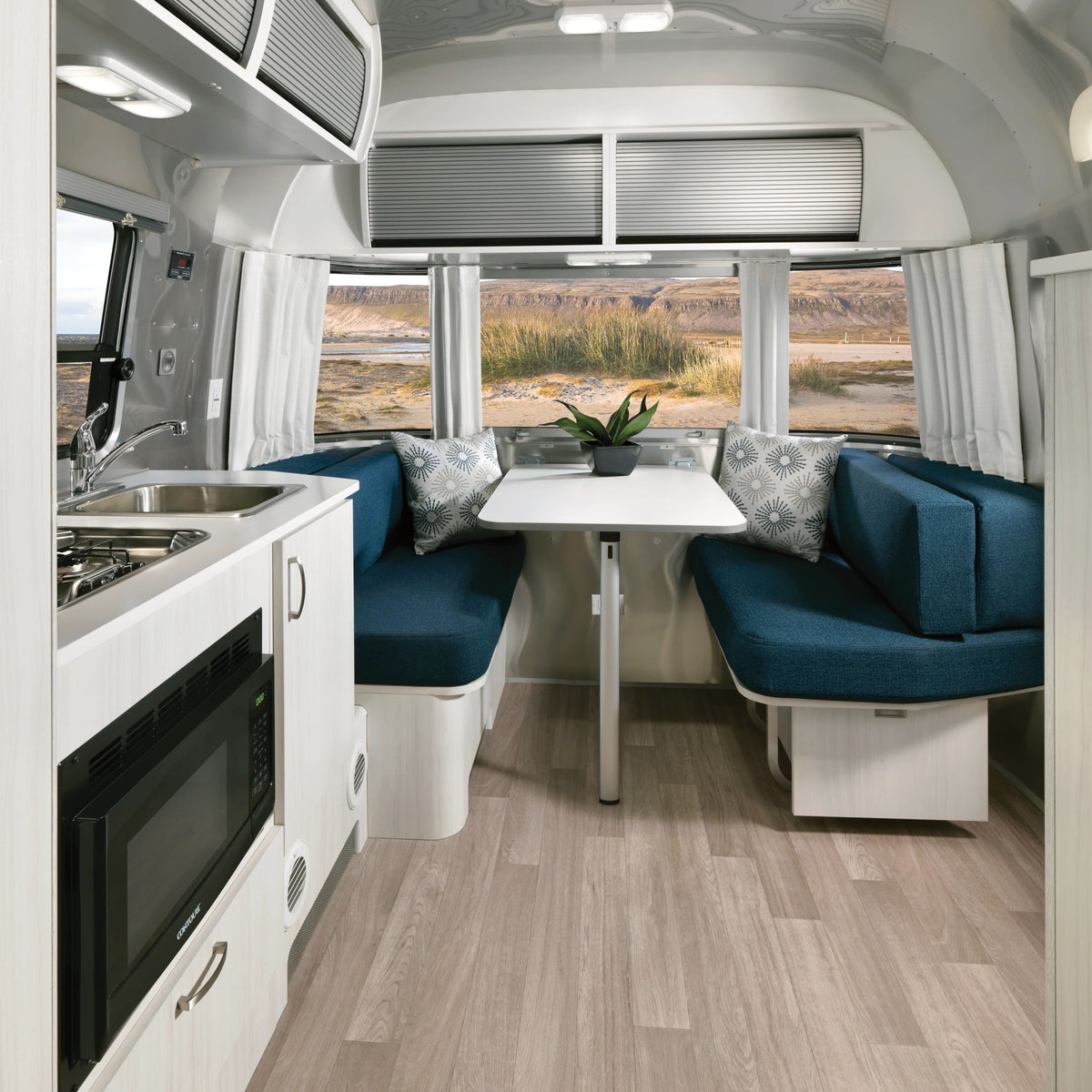 A modern RV interior with a kitchenette, microwave, and dining area featuring blue cushioned seats, a white table, and large windows adorned with Airstream Solid Custom Curtains for Bambi Travel Trailers overlooking a scenic landscape.