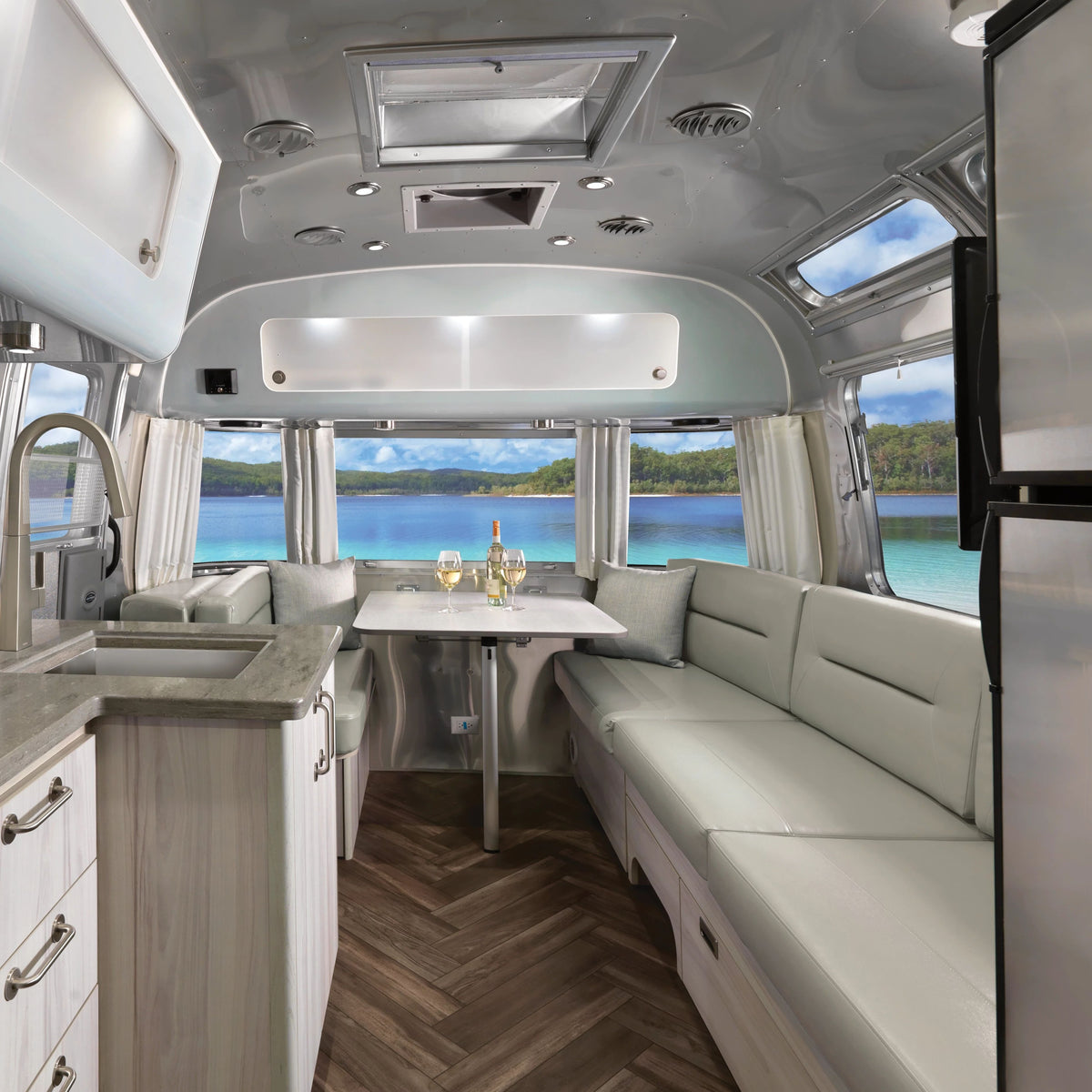 The interior of a modern RV features cream seating, a dining table set for four, a kitchen area, and large windows adorned with Airstream Solid Custom Curtains for International Travel Trailers, framing views of a lake and trees outside on a sunny day.