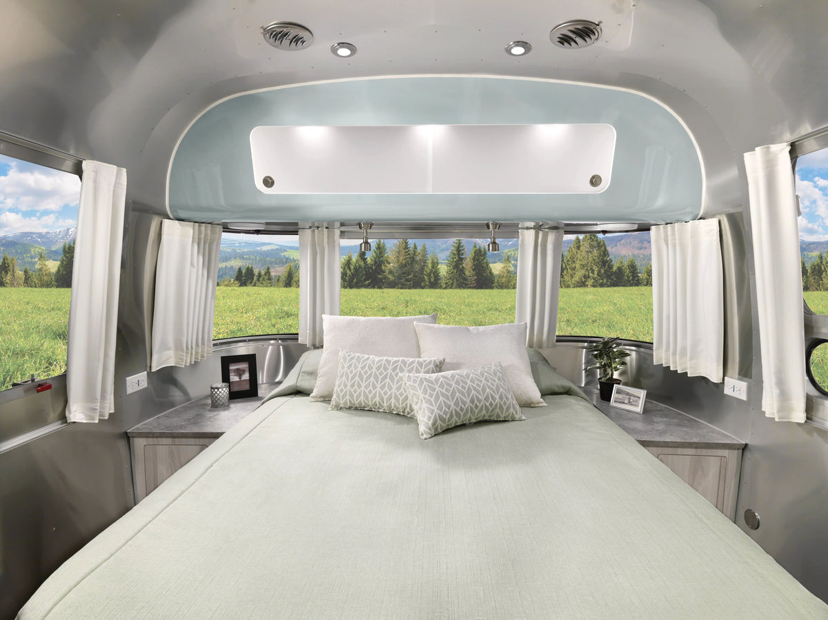 A bed with light green bedding inside a camper overlooks fields and trees through expansive windows dressed in Airstream Solid Custom Curtains for International Travel Trailers in crisp white. Pillows, framed photos, and a small plant accentuate the side tables, enhancing the cozy ambiance—perfect for those seeking chic replacement curtains or stylish Airstream curtains.