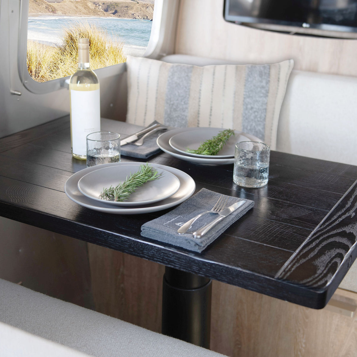 A black table set for two features white plates from the Airstream x Pottery Barn Dinnerware 16 Pc Set, accompanied by glasses of water, silverware, a bottle of white wine, and sprigs of rosemary. The cozy space includes striped pillows and a window revealing a scenic outdoor view.