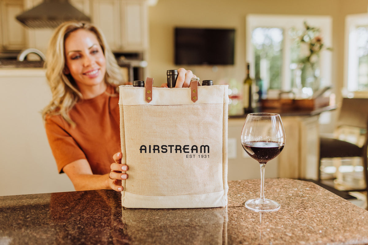 A woman smiles while holding an Airstream Insulated 3 Bottle Wine Bag filled with wine bottles on a kitchen counter next to a glass of red wine.
