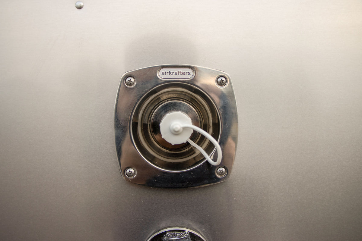 Close-up of a metal circular fuel cap labeled nLet™ by Airkrafters, attached to a smooth metallic surface and secured with a white tether—reminiscent of the stainless steel city water inlet found on an Airstream travel trailer.