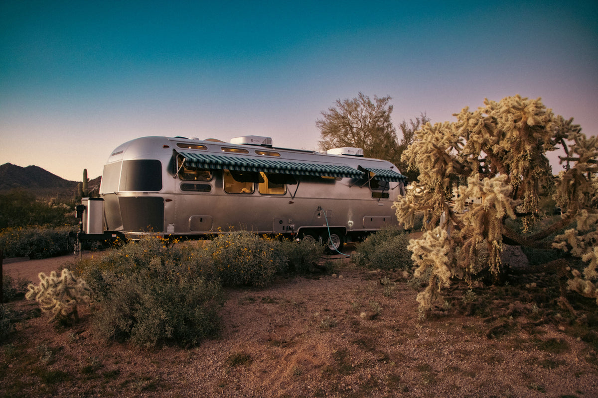 A silver Airstream trailer with a green striped awning is parked in a desert landscape at sunset, surrounded by cacti and bushes, with distant mountains and a clear sky in the background.