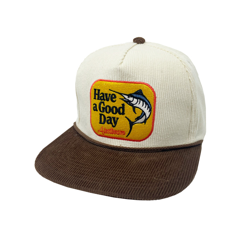 The Airstream Have a Good Day Corduroy Trucker Hat is a cream and brown baseball cap featuring a yellow patch on the front with a fish and the cheerful text “Have a Good Day.”.