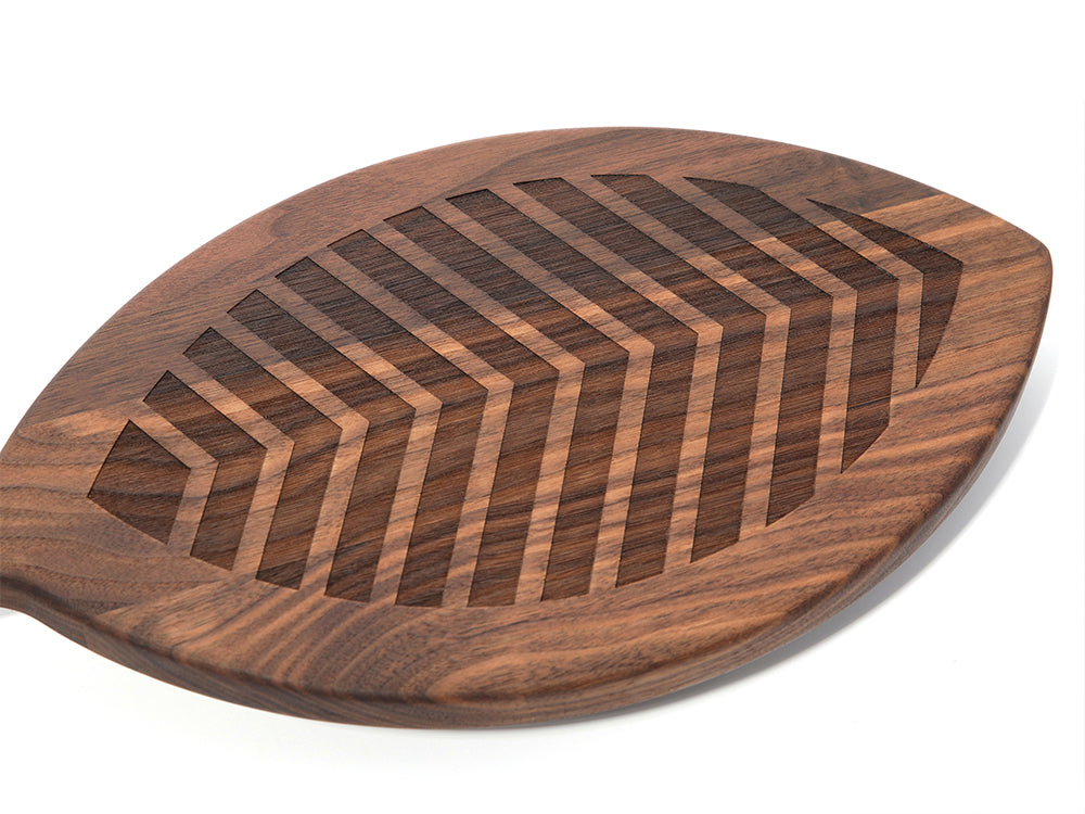 The Frank Lloyd Wright x Airstream Charcuterie Board is a walnut serving piece shaped like a leaf, featuring carved geometric lines that mimic leaf veins, and is shown on a white background. This board blends natural elegance with design details inspired by both Airstream and Frank Lloyd Wright.