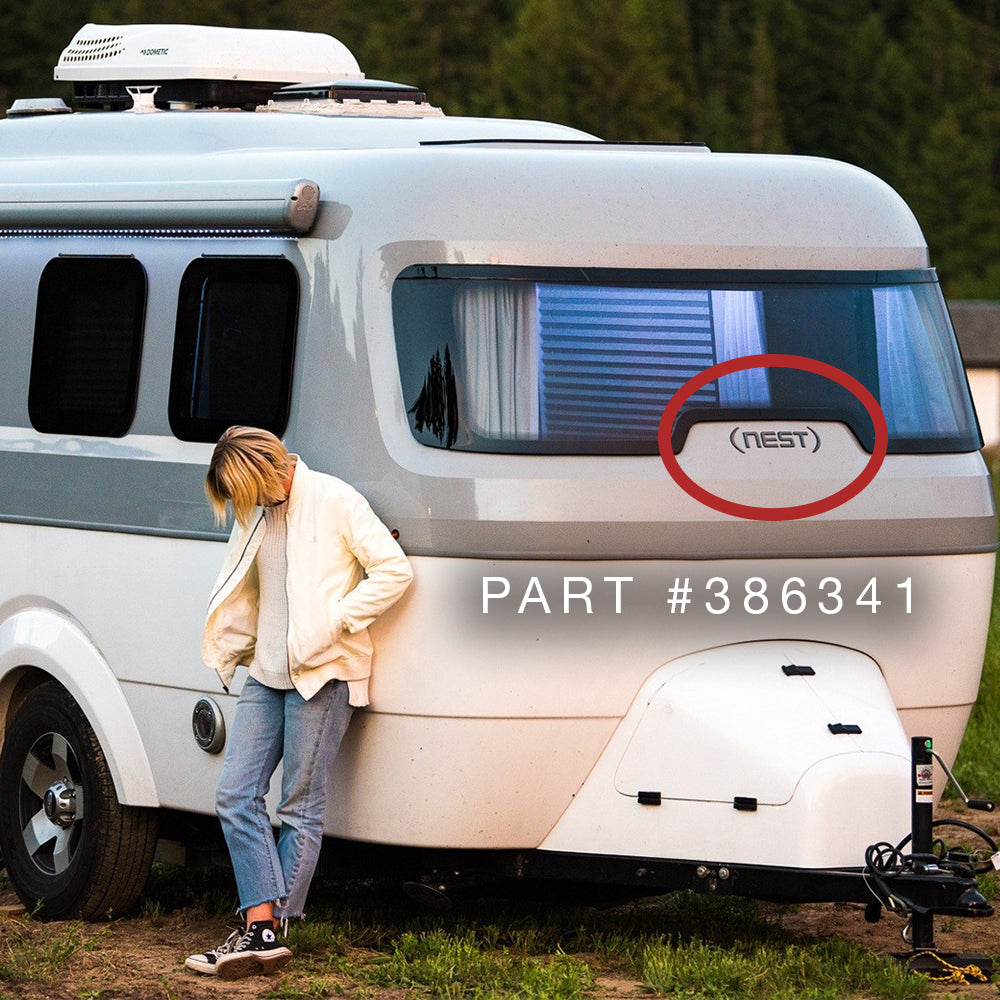 A person in a white jacket leans against a small gray and white camper trailer featuring a window with blinds and an NLA Airstream Nest Black Chrome Logo Decal (Part #386341), with the word “NEST” highlighted by a red circle.